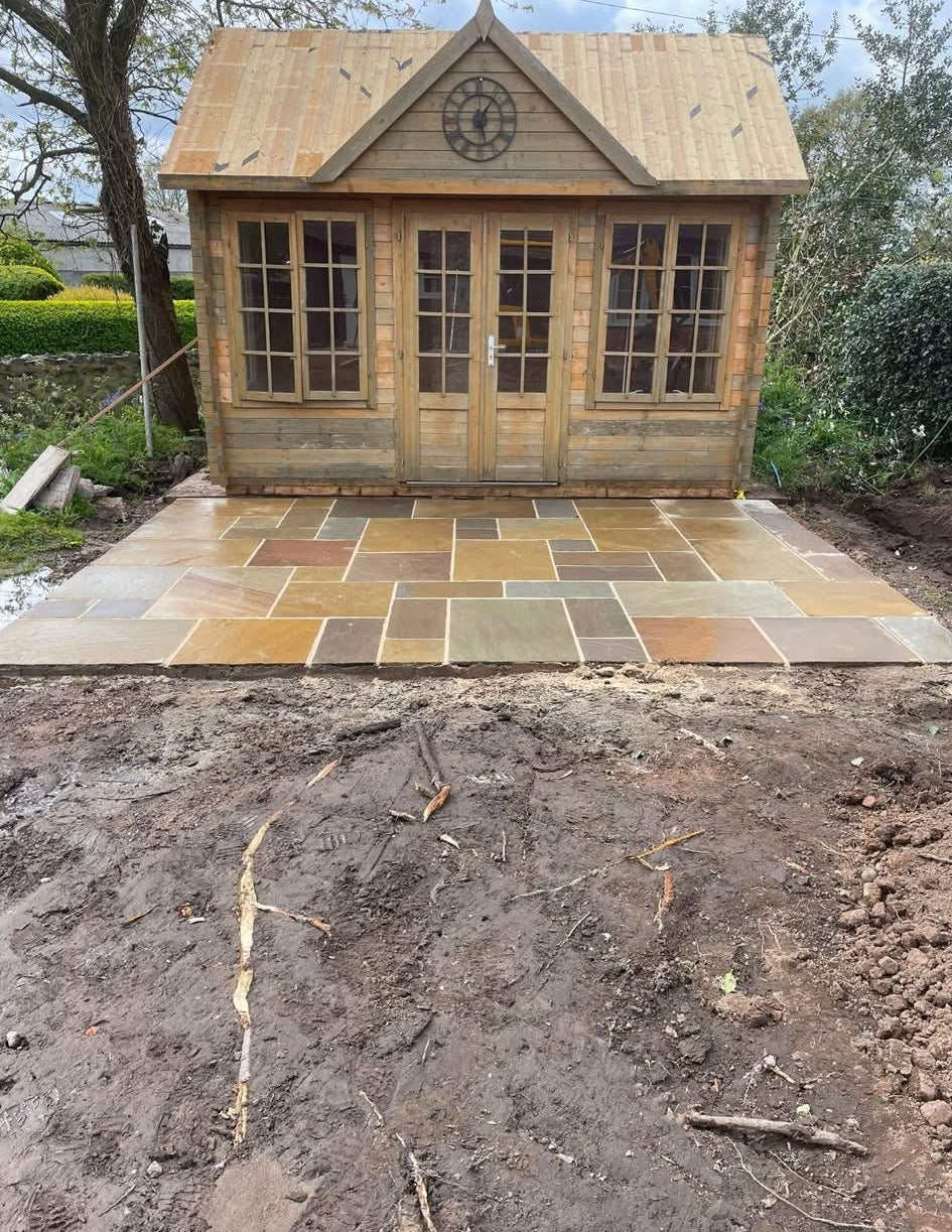 A small wooden garden shed with a pitched roof, multi-paned windows, and double doors, set outdoors on a stone patio with a muddy yard in the foreground.